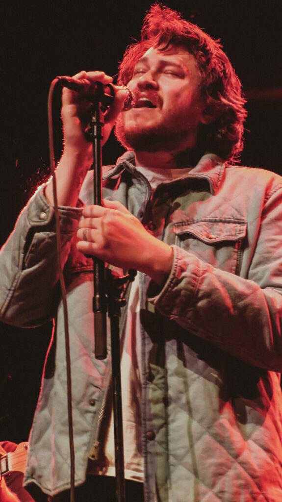 A live shot from Braison Cyrus’s Austin concert performance with guitar, captured in warm colors and a calm, folk-inspired setting.