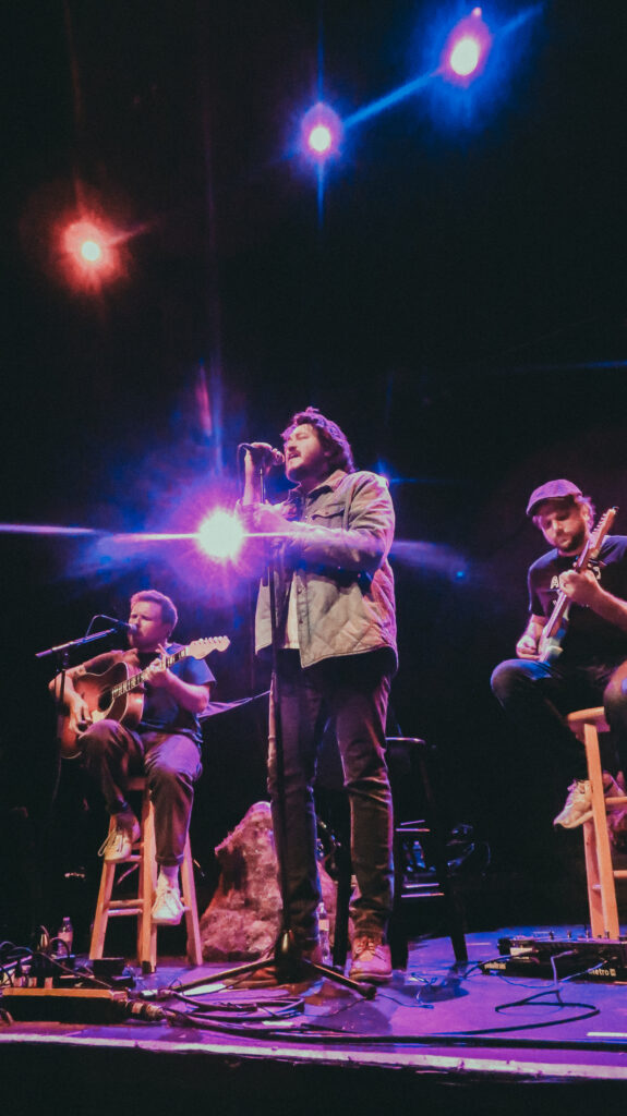 A live shot from Braison Cyrus’s Austin concert performance with guitar, captured in warm colors and a calm, folk-inspired setting.
